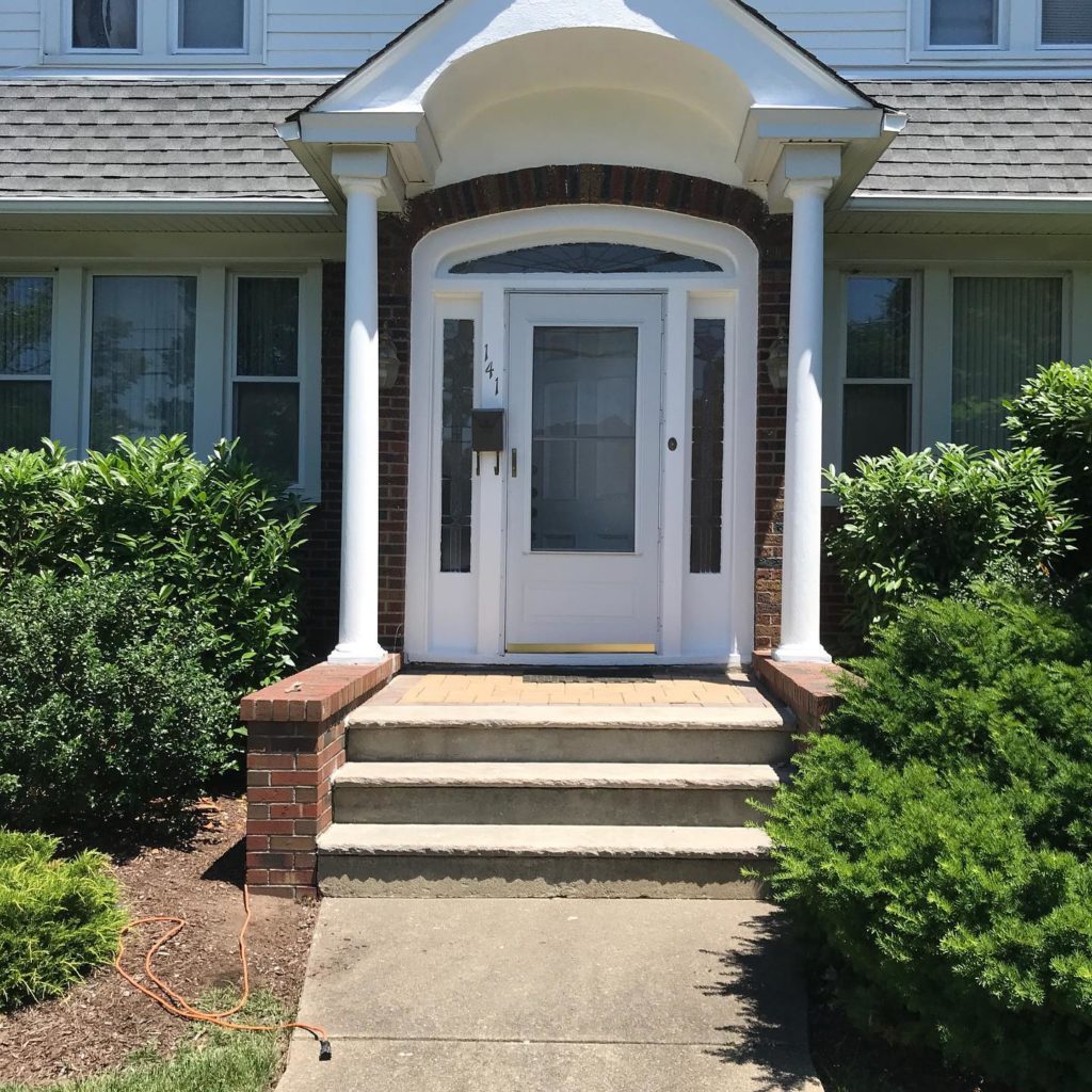 Front entrance of a house with white storm door, brick steps, white columns, and surrounding shrubs.